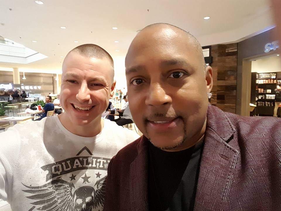 Chance Trahan (left, white graphic t-shirt, smiling) taking a close selfie with entrepreneur Daymond John (right, maroon blazer) indoors at a mall or food court.