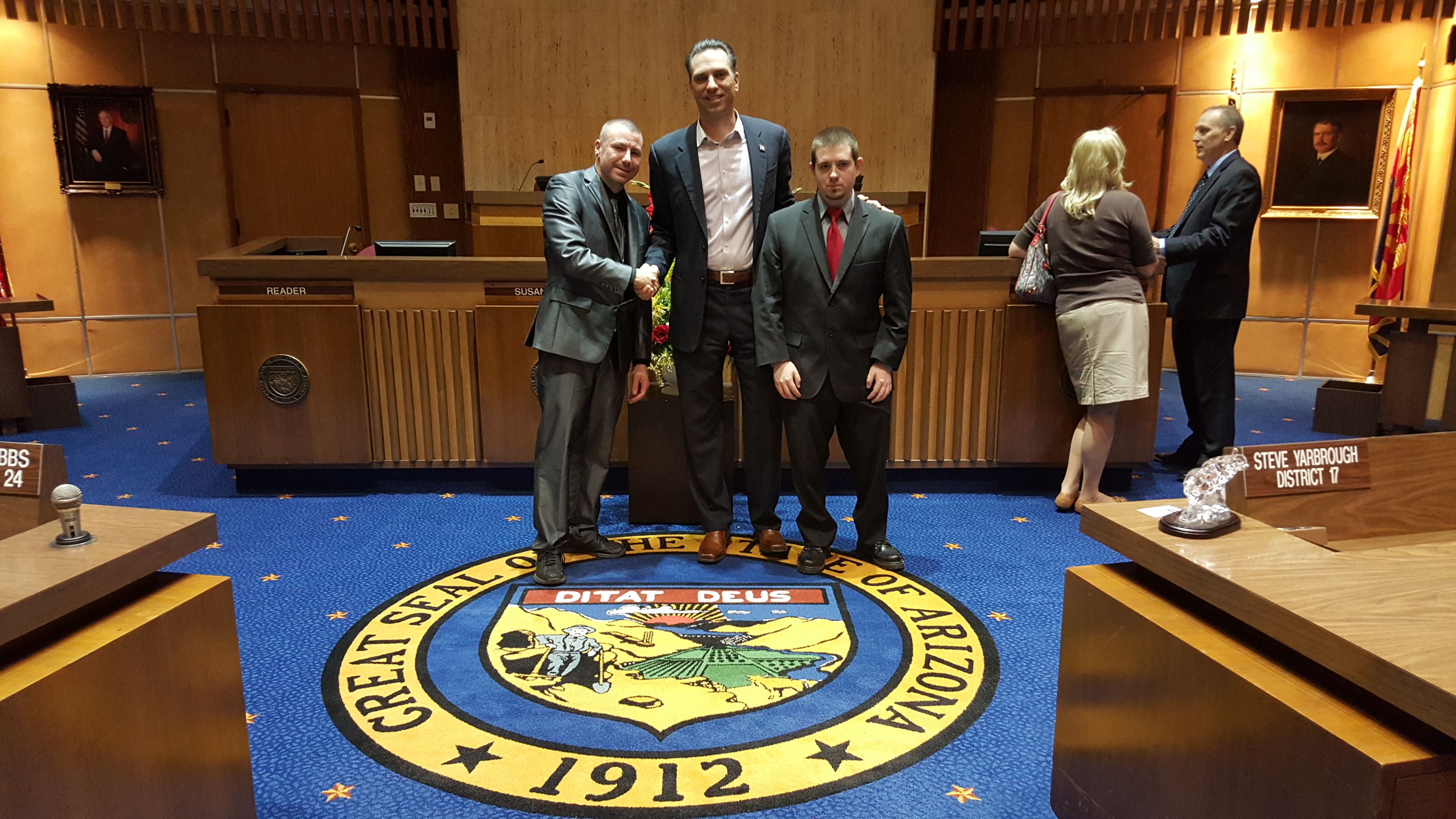 chance trahan is wearing a silver suit and is shaking hands with politician steve smith in the arizona senate chamber, craig brittain is to the right, they are standing in front of the great seal of arizona